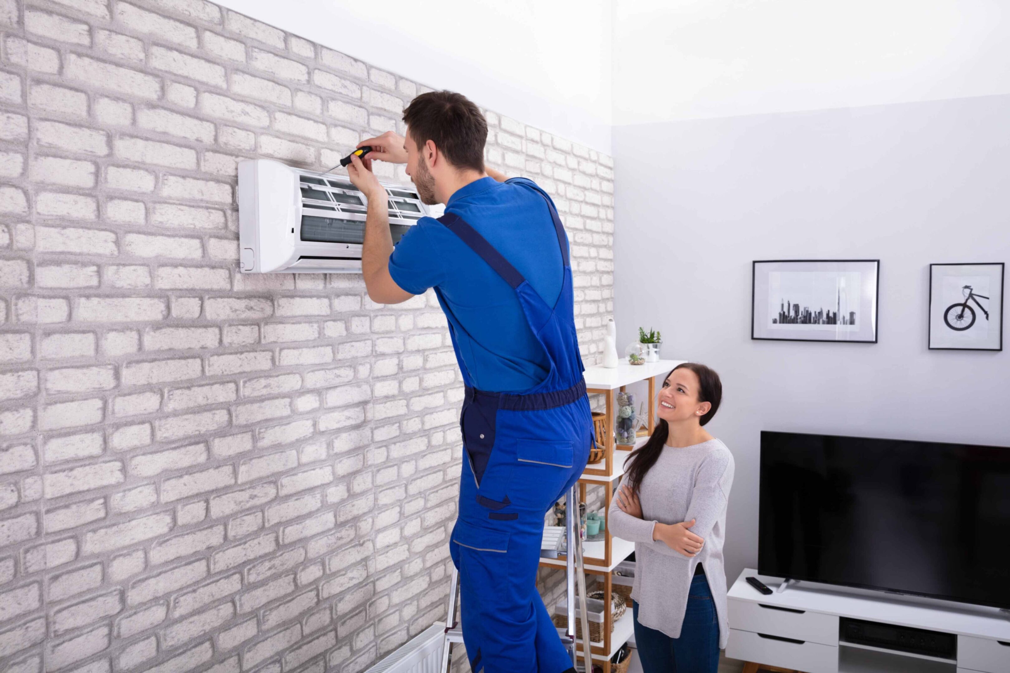 A technician in blue overalls repairs a wall-mounted air conditioner while a woman stands nearby watching in a modern living room.