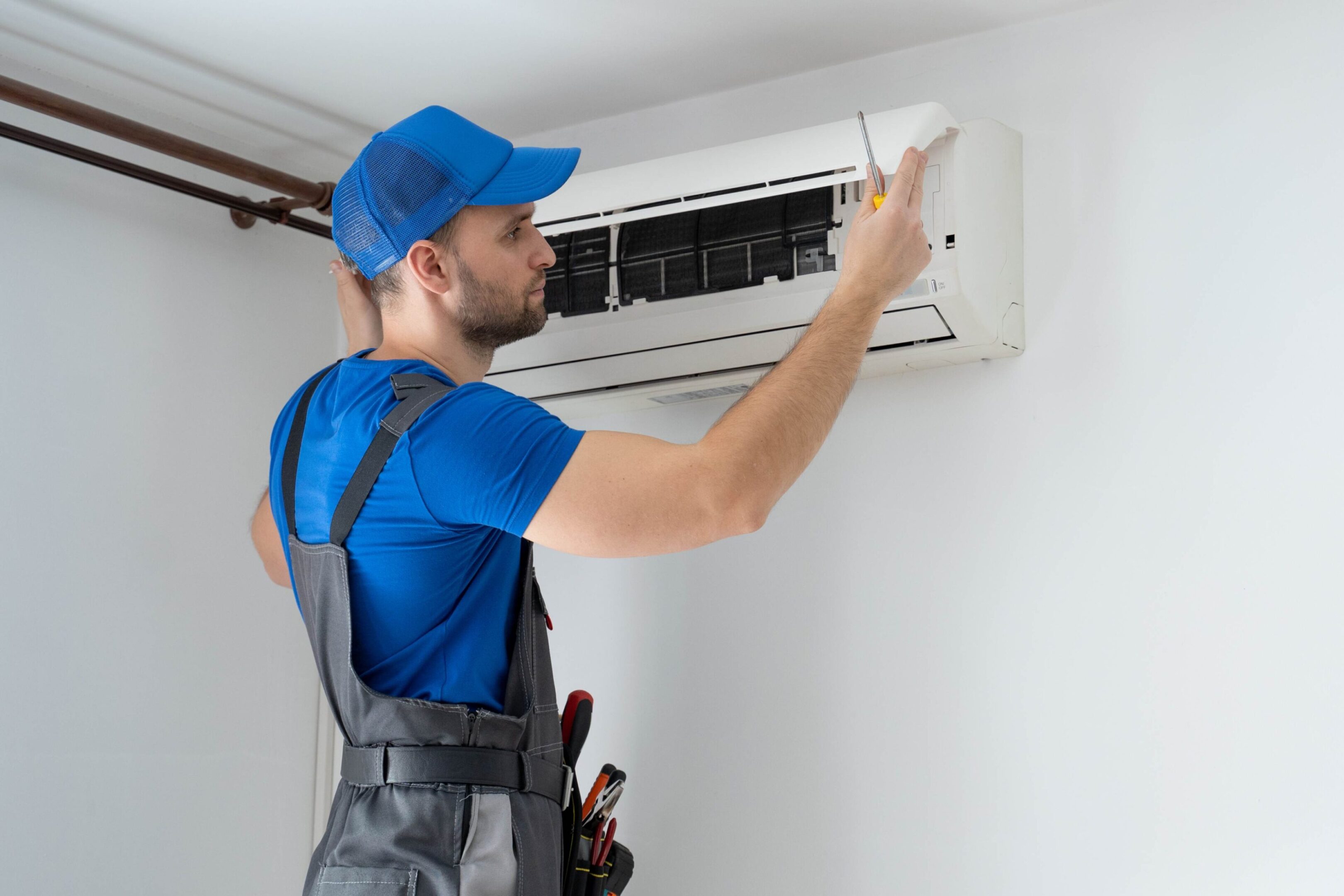A technician in blue uniform and cap is repairing a wall-mounted air conditioner with a screwdriver.