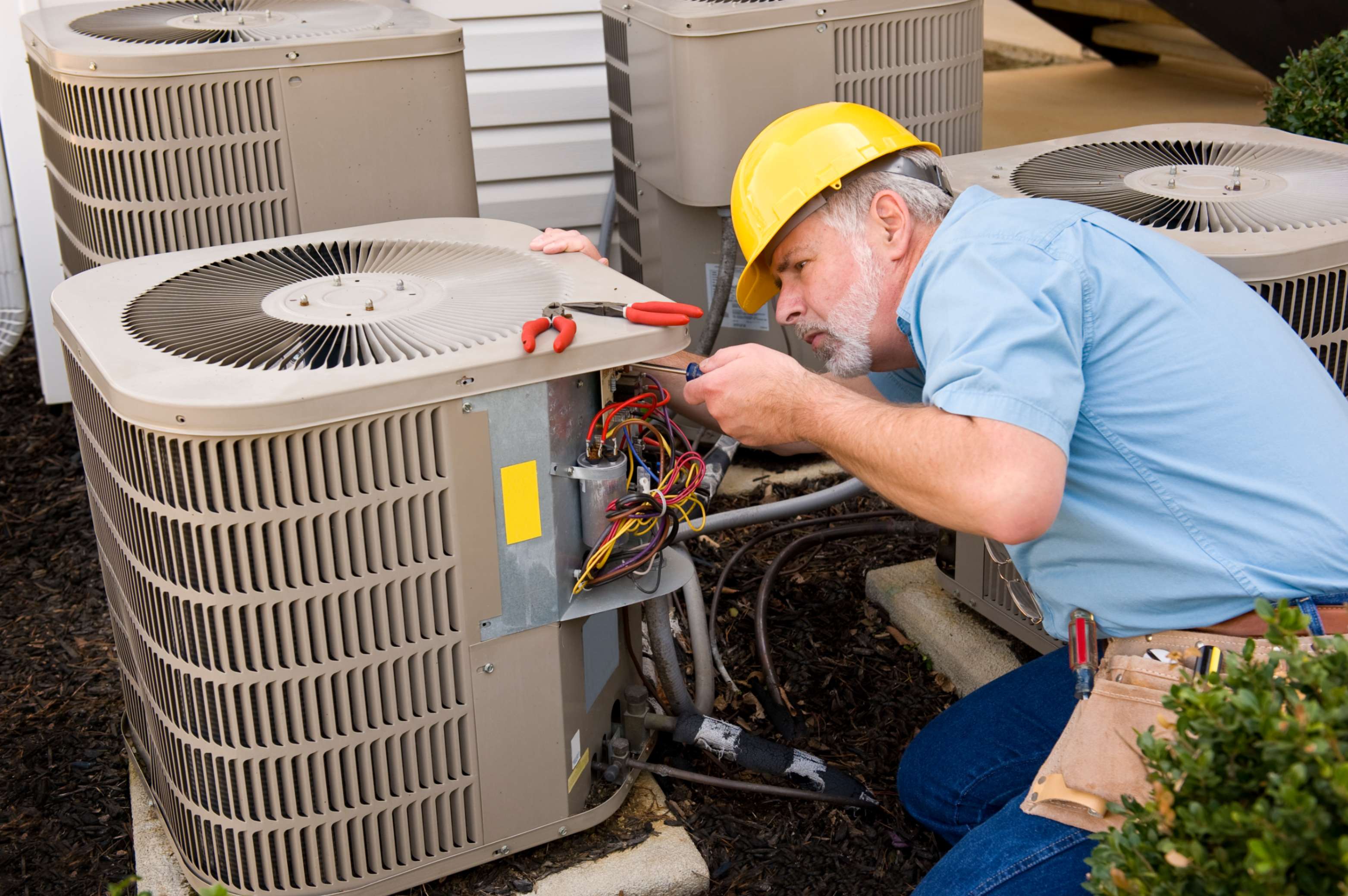 A technician wearing a yellow hard hat and tool belt repairs the wiring inside an outdoor air conditioning unit next to a building.